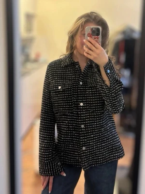 Svart och vit overshirt med knappar - Säljer en svart overshirt med vita ränder och silverfärgade knappar. Overshirten har två bröstfickor med lock och klassisk krage. Materialet ser ut att vara tjockt och strukturerat, perfekt för lager-på-lager. Passar dig som gillar en avslappnad och trendig look.