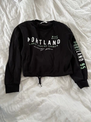 Svart cropped hoodie - Svart cropped hoodie från H&M med trycket 'PORTLAND WEST HILLS' framtill och 'PORTLAND 95' på ena ärmen. Tröjan har långa ärmar, ribbade muddar och dragsko i nederkanten. Perfekt för en avslappnad streetstyle-look.🤍