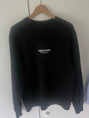 Svart sweatshirt från Jack & Jones - Snygg svart sweatshirt från Jack & Jones Originals med tryck 'Originals Studio' på bröstet. Klassisk rund halsringning och långa ärmar. Perfekt för en clean och enkel stil.