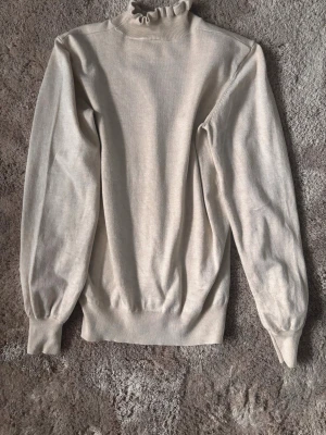 Beige stickad tröja med volangkrage - Säljer en beige stickad tröja med lång ärm och volangdetalj i halsen. Tröjan har ribbade muddar vid ärmslut och nederkant, samt en mjuk och följsam passform. Perfekt för dig som gillar stilrena och trendiga plagg.