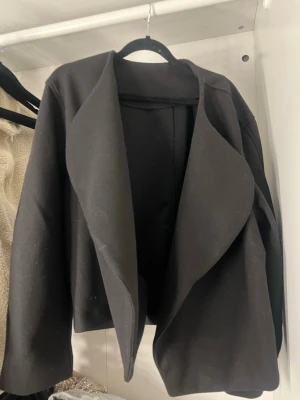 Svart kappa med öppen front - Stilren svart kappa med öppen front och breda slag. Modellen är kortare och har en avslappnad passform med långa ärmar. Perfekt för lager-på-lager och enkel att matcha med olika outfits. Materialet känns mjukt och har en lätt struktur.