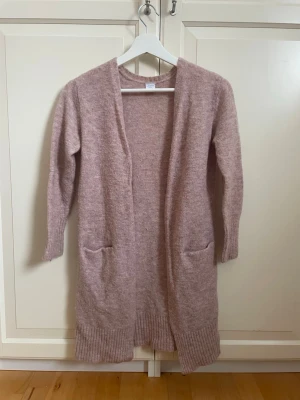 Ljusrosa lång kofta med fickor - Mysig ljusrosa lång kofta med öppen front och två praktiska fickor framtill. Koftan har ribbstickade muddar och nederkant, vilket ger en snygg detalj. Perfekt att slänga på sig över en t-shirt eller blus. Materialet är mjukt och ger en avslappnad look.