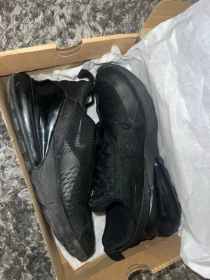 Nike Air Max 270 helsvarta sneakers - Säljer ett par helsvarta Nike Air Max 270 sneakers med stor synlig Air-enhet i hälen. Ovandelen är i mesh och syntetmaterial för bra andningsförmåga och komfort. Klassisk Nike Swoosh på sidan och modern design som passar till streetwear.Litet hål vid tån men svårt att märka , kan också fixas lätt , tappat bort 1 snörre !