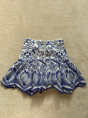 Mönstrad volangkjol från Zara - Säljer en kort kjol med volang och vackert blått mönster på vit botten. Kjolen har knappar framtill, samt ett unikt och detaljerat print som verkligen sticker ut. Perfekt för dig som gillar att synas och vill ha något speciellt i garderoben. 🩵