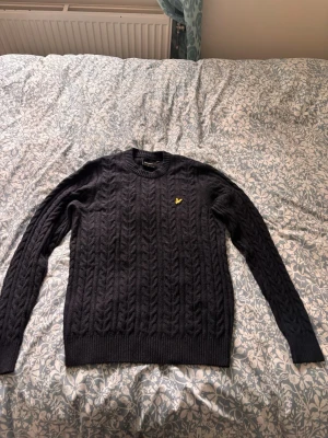 Svart kabelstickad tröja Lyle & Scott - Kabelstickad svart tröja från Lyle & Scott med rund hals och långa ärmar. Tröjan har ett gult broderat logomärke på bröstet och ribbade muddar vid ärmslut och nederkant. Finns även lite mörkblå nyans, men skulle säga att den är svart. Kostar ungefär 1200kr ny