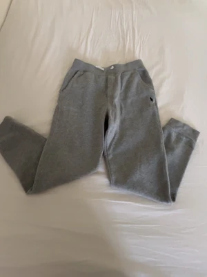 Grå mjukisbyxor från Polo Ralph Lauren - Gråa mjukisbyxor från Polo Ralph Lauren med klassisk passform och resår i midjan. Byxorna har sidofickor och en liten broderad logga på vänster sida. Tillverkade i mjukt och bekvämt material, perfekta för chill dagar.