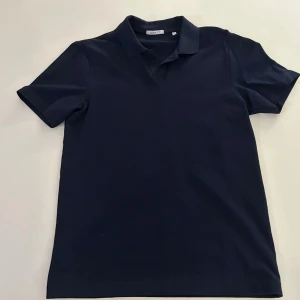 Mörkblå pikétröja - En stilren mörkblå pikétröja med klassisk krage och korta ärmar. Perfekt till jeans eller shorts för en avslappnad men snygg look. Enkel design utan mönster eller detaljer.