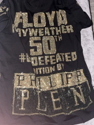 Svart Philipp Plein t-shirt med guldtext - Säljer en svart t-shirt från Philipp Plein med stora glittriga guldbokstäver och detaljer framtill. Texten hyllar Floyd Mayweather och har en exklusiv känsla med paljetter och bling. T-shirten har korta ärmar och rund hals.