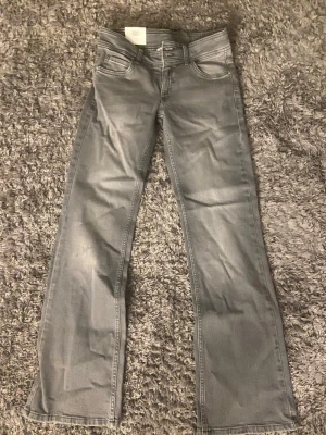 Grå bootcut jeans från Kappahl - Snygga grå bootcut jeans från Kappahl med låg midja och klassiska fem fickor. Jeansen har en lätt utsvängd passform nedtill och är tillverkade i mjukt denimtyg. Perfekta för en avslappnad och trendig look.