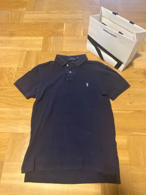 Mörkblå pikétröja från Ralph Lauren - Klassisk mörkblå pikétröja från Polo Ralph Lauren med korta ärmar och krage. Tröjan har två knappar vid halsen och den ikoniska broderade loggan på bröstet. Tillverkad i mjuk bomull för en bekväm känsla.