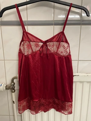 Rött nattlinne med spetsdetaljer - Snyggt rött nattlinne med tunna axelband och transparent spets upptill samt nertill. Dekorerat med blommönstrad spets och en liten rosett framtill. Materialet är glansigt och mjukt, perfekt för en bekväm kväll hemma. Står att den är i storlek L men är mer som en S.