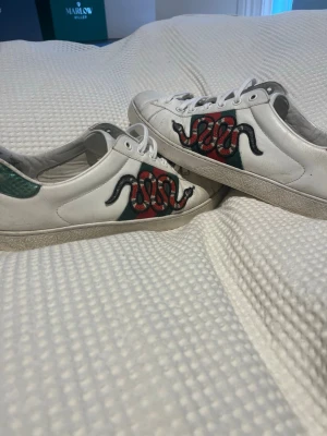 Gucci Ace sneakers med orm - Säljer ett par vita Gucci Ace sneakers med den ikoniska broderade ormen på sidan, gröna och röda detaljer samt vita skosnören. Skorna har låg profil och klassisk siluett, tillverkade i läder med gummisula. Perfekta för dig som vill sticka ut med lyxig streetstyle. Finns tyvärr ingen box med