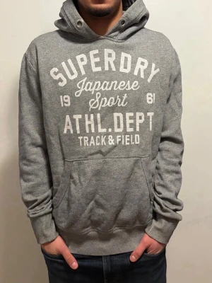 Extremt sällsynt Grå Superdry hoodie med tryck - Snygg grå hoodie från Superdry med stort vitt tryck på bröstet där det står 'Superdry Japanese Sport ATHL. DEPT Track & Field'. Hoodien har huva med dragsko och en klassisk magficka. Perfekt för en avslappnad och sportig stil.