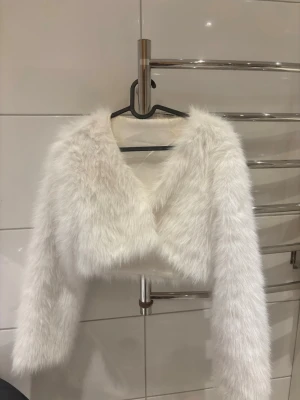 Vit kort pälsjacka med lång ärm - Supermjuk och fluffig vit pälsjacka i kort modell med långa ärmar. Jackan har en öppen front och är tillverkad i fuskpäls som ger en lyxig känsla. Perfekt för att lyfta din outfit med en trendig och cool vibe.
