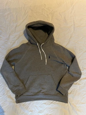 Grå hoodie från Polo Ralph Lauren - Snygg grå hoodie från Polo Ralph Lauren med vit dragsko i huvan och klassisk känguruficka. Liten mörkblå logga broderad på bröstet. Tillverkad i mjuk bomullsblandning, perfekt för en avslappnad och stilren look.