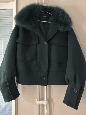 Mörkgrön pälsjacka från Meotine - Stilren mörkgrön pälsjacka från Meotine med fluffig fuskpälskrage, stora fickor framtill och markerade knappar. Jackan har en boxig, kortare passform och breda ärmslut med knappdetaljer. Perfekt för att hålla värmen och se trendig ut under vintern.