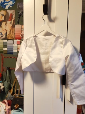 Vit bolero - Säljer en vit bolero med långa ärmar och öppen front. Tillverkad i ett slätt, vävt tyg som ger en elegant känsla. Perfekt att slänga över en klänning eller topp för en extra touch. Enkel och stilren design utan synliga detaljer eller mönster.