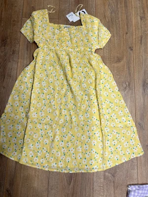 Gul blommig klänning med kort ärm - Söt gul klänning med vita och gröna blommor, fyrkantig ringning och korta ärmar. Klänningen har en lätt utställd kjol och är perfekt för varma dagar. Tillverkad i bomull för en luftig känsla. Nypris på den är 349kr!