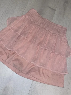 Rosa volangkjol med prickigt mönster - Supersöt kort kjol i ljusrosa med volanger och diskret prickigt mönster. Kjolen har smock i midjan för en bekväm passform och är gjord i ett lätt, luftigt material som känns perfekt för varmare dagar.