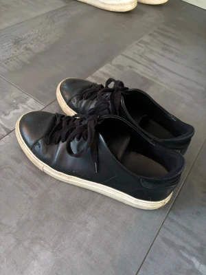 Svarta låga sneakers i läder - Stilrena svarta sneakers i läder med svart snörning och vit sula. Klassisk låg modell som passar till det mesta och ger en clean look. Perfekta för dig som gillar minimalistisk stil.