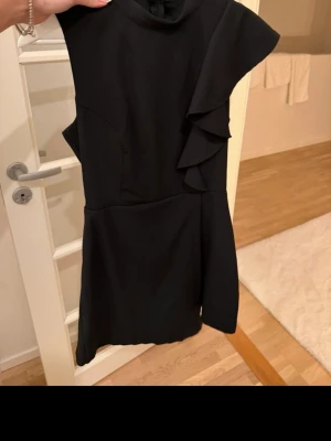 Svart kort volangklänning - Snygg svart kort klänning med volangdetaljer på axeln och sidan. Ärmlös design och figurnära passform som ger en elegant siluett. Perfekt för dig som vill ha en stilren look med det lilla extra. Dragkedja i ryggen för enkel påklädning.