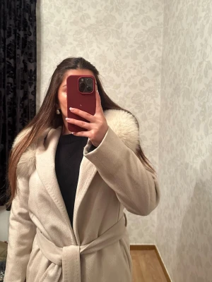 Beige kappa från hollies - Säljer en stilren beige kappa från hollies och knytskärp i midjan. Kappan har en klassisk design med långa ärmar och en mjuk, lyxig känsla. Perfekt för att hålla sig varm under kalla dagar och ge en elegant look.