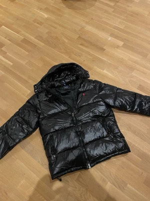 Svart pufferjacka från Polo Ralph Lauren - Säljer en svart pufferjacka från Polo Ralph Lauren med blank finish och röd logga på bröstet. Jackan har huva, dragkedja framtill och praktiska fickor. Materialet är syntet och jackan är perfekt för kalla vinterdagar.