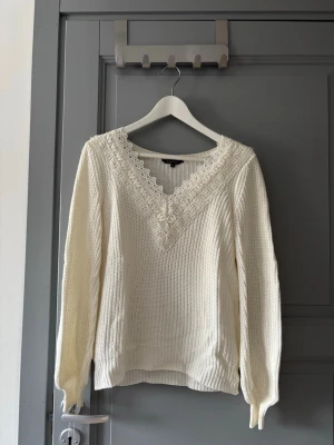 Stickad tröja  - Säljer en vit stickad tröja från Vero Moda med v-ringning och dekorativa spetsdetaljer runt halsen. Tröjan har långa ärmar och ribbade muddar vid ärmslut och nederkant. Perfekt för både vardag och fest. 
