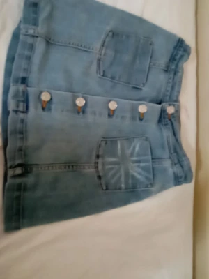 Blå jeanskjol med knappar - Säljer en blå jeanskjol från Your Denim med stora framfickor och snygga silverfärgade knappar längs hela framsidan. Kjolen har en klassisk rak passform och är gjord i slitstarkt denimtyg. Perfekt för dig som gillar en enkel men trendig stil.