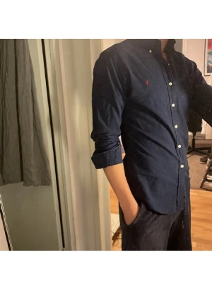 Mörkblå skjorta från Ralph Lauren - Klassisk mörkblå skjorta från Ralph Lauren med button-down krage och vita knappar. Skjortan har lång ärm och den ikoniska röda logotypen broderad på bröstet. Tillverkad i mjuk bomull för en bekväm känsla och snygg passform.