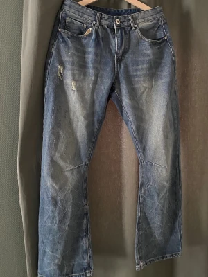 Bootcut jeans med schyssta slitningar - Säljer ett par blå jeans med raka ben och slitna detaljer på låret. Jeansen har klassisk femficksdesign och dragkedja med knapp. Perfekta för en avslappnad och trendig look.