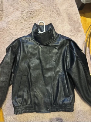 Svart skinnjacka med hög krage - Säljer en svart skinnjacka med hög krage och asymmetrisk knäppning. Jackan har två snedställda fickor framtill och resår i midjan för en schysst passform. Perfekt för dig som vill ha en stilren och edgy look.