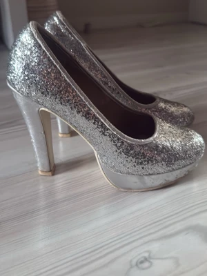 Glittriga silvriga pumps från DinSko - Säljer ett par silvriga pumps från DinSko med glittrig yta och hög platåklack. Skorna har rundad tå och är täckta av silverglitter som verkligen drar blickarna till sig. Perfekta för dig som vill sticka ut med din stil.