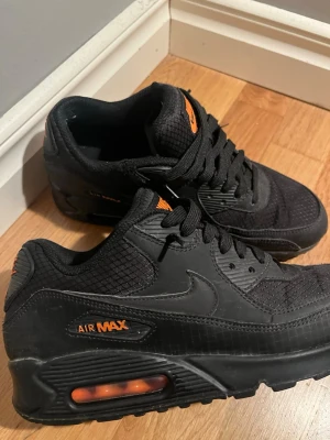 Nike Air Max 90 svart/orange sneakers - Snygga Nike Air Max 90 sneakers i svart med orange detaljer. Skorna har en klassisk låg siluett, mesh och syntetmaterial på ovandelen samt den ikoniska Air Max-dämpningen i sulan. Perfekta för dig som gillar streetwear och sportig stil.