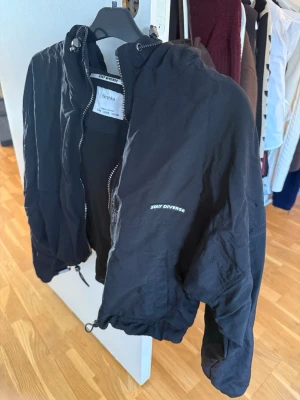 Svart vindjacka från Bershka - Svart vindjacka från Bershka med huva och dragkedja framtill. Jackan har snörning i nederkant, meshfoder och texten 'STAY DIVERSE' på bröstet. Perfekt för blåsiga dagar och enkel att matcha med olika outfits. Med reflex på baksidan