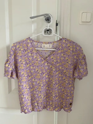 Lila blommig omlottopp från UO - Supersöt lila omlottopp från Urban Outfitters med små gula och vita blommor. Toppen har korta puffärmar, v-ringning och volangkant nedtill. Perfekt för dig som vill ha en färgglad och somrig vibe i garderoben!