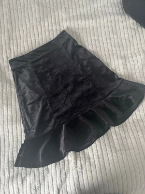 Svart sammetskjol med volang - Supersnygg svart kortkjol i mjuk sammet med volangkant nedtill och dragkedja baktill. Kjolen har en lätt glansig finish och sitter tajt upptill för en modern look. Perfekt att styla med en croppad topp eller stickad tröja.