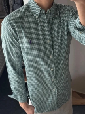 Grön rutig skjorta från Ralph Lauren - Säljer en grön och vit rutig skjorta från Ralph Lauren med klassisk button-down krage och lila broderad logga på bröstet. Skjortan har långa ärmar och vita knappar framtill. Perfekt för dig som gillar stilrena och tidlösa plagg.