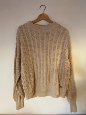 Pernilla Wahlgren kabelstickad tröja S-L - Snygg beige kabelstickad tröja med bred ribbad krage och muddar. Tröjan har ett klassiskt mönster med vertikala flätor och en liten gulddetalj vid nederkanten. Perfekt för lager på lager och riktigt mysig. Från Pernilla Wahlgrens kollektion. Märkt med storlek L men upplevs mer som en M eller oversized S. Knappt använd, jättefint skick. 