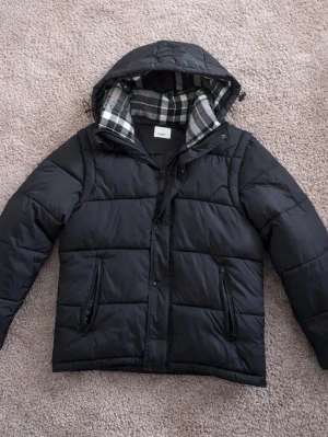 burberry jacka - Snygg svart pufferjacka med huva och dragkedja framtill. Jackan har två dragkedjefickor och ett rutigt foder i svart, vitt och rött. Perfekt för kalla dagar och ger en avslappnad streetwear-look. Materialet är quiltat och jackan har en klassisk passform.