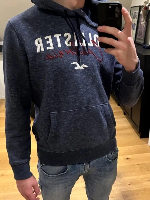 Mörkblå Hollister hoodie - Snygg mörkblå hoodie från Hollister med vit och röd text på bröstet där det står 'Hollister California'. Hoodien har en stor magficka, dragsko i huvan och ribbade muddar vid ärmar och nederkant. Perfekt för en avslappnad och cool stil.