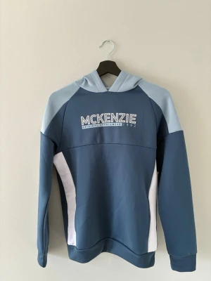 Blå hoodie från McKenzie - Snygg hoodie från McKenzie i blått med ljusblå ärmar och vita sidopaneler. Stor logga på bröstet med texten 'McKenzie Original Sportswear 1992'. Hoodien har huva och är tillverkad i mjukt bomullsmaterial. Perfekt för en avslappnad streetwear-look.