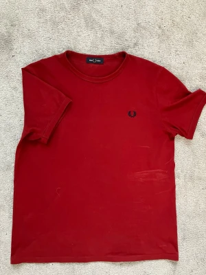 Röd t-shirt från Fred Perry - Snygg röd t-shirt från Fred Perry med klassisk rund hals och diskret logga broderad på bröstet. Tillverkad i mjuk bomull och har en normal passform. Perfekt till jeans eller shorts för en enkel och stilren look.