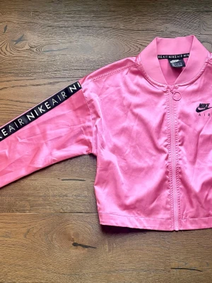  Nike Air  - Snygg rosa bomberjacka från Nike Air med dragkedja framtill och ribbad krage. Jackan har svart Nike Air-tryck på bröstet och svart band med text längs ena ärmen. Perfekt för dig som vill sticka ut med sportig stil.