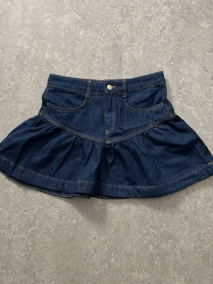 Mörkblå jeanskjol med volang - Trendig mörkblå jeanskjol med volangkant och klassiska fickor fram. Kjolen har en kort modell och stängs med knapp och dragkedja. Perfekt för dig som gillar denim och vill ha en cool look.