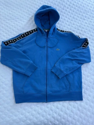 Blå hoodie från Lacoste med dragkedja - Snygg blå hoodie från Lacoste med dragkedja framtill och huva. Har svarta band med Lacoste-logga längs ärmarna och den klassiska krokodilloggan på bröstet. Tillverkad i mjukt bomullsmaterial och har fickor på sidorna. Perfekt för en avslappnad streetwear-look. Storlek XL men passar även M. Vad jag vet så säljs inte denna tröjan längre, köpt på Zalando för 2100kr.