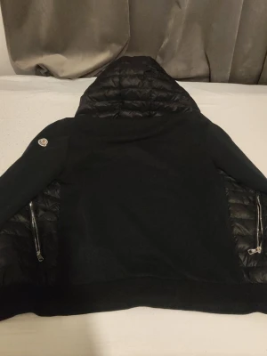 Svart dunjacka med huva från Moncler - Snygg svart dunjacka från Moncler med huva och quiltade partier. Jackan har två dragkedjeförsedda fickor och en Moncler-logga på ärmen. Materialet är en mix av dun och textil, vilket ger både värme och stil. Perfekt för kalla vinterdagar.