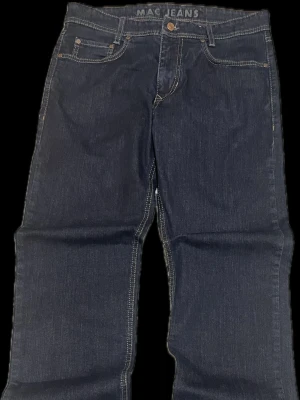 Mörkblå raka jeans från MAC Jeans - Snygga mörkblå jeans från MAC Jeans med klassisk femficksmodell och raka ben. Jeansen har kontrastsömmar och metallknapp i midjan. Perfekta för dig som gillar en tidlös och stilren look. Materialet är denim i bomull.
