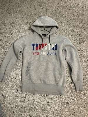Grå hoodie från Trapstar  - Snygg grå hoodie från Trapstar med huva och känguruficka. Framsidan har ett stort tryck i blått, rött och vitt med texten 'Trapstar It’s a Secret'. Tillverkad i mjukt bomullsmaterial och har svarta dragsnören vid huvan.