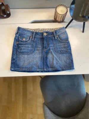 Blå jeanskjol med fickor - Snygg blå jeanskjol i kort modell med stora framfickor och bakfickor med knappdetaljer. Kjolen har bälteshällor och klassisk knappstängning framtill. Perfekt för dig som gillar en avslappnad och trendig stil. Midjemåttet är 34 cm🩷